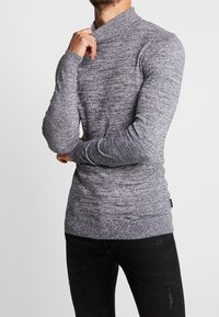 Pull gris en maille avec un col montant et des manches longues, présentant un motif texturé et une silhouette ajustée. Associé à un jean foncé.