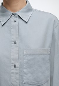 Ανοιχτό γκρι πουκάμισο με κουμπιά και μυτερό γιακά, με τσέπη στο στήθος, φορεμένο από ένα άτομο σε απλό φόντο.