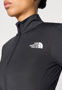 Parte superior deportiva negra con cremallera de un cuarto, hecha de tela suave, con un cuello alto y firmada con un logotipo blanco de "The North Face" en el pecho.