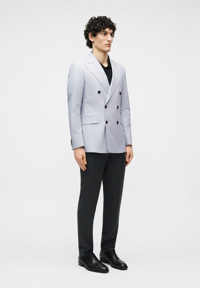 Άνδρας που στέκεται φορώντας ανοιχτογκρι διπλόπατο σακάκι, μαύρο πουκάμισο, μαύρο παντελόνι και μαύρα παπούτσια φόρμας μπροστά σε απλό φόντο.