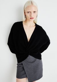 Pulover negru, supradimensionat, din tricot, cu un design specific în față, asortat cu o fustă încrețită de culoare gri, cu model herringbone. Textură netedă în ansamblu.