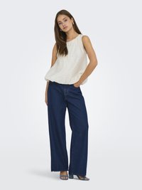 Un haut sans manches beige clair associé à un jean en denim bleu foncé à jambes larges. Le mannequin se tient les mains dans les poches, portant des sandales à talons bleues.