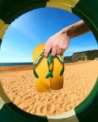 Infradito gialle con cinturini verdi e suole testurizzate sono tenute da una mano, viste attraverso una cornice circolare contro uno sfondo di spiaggia.