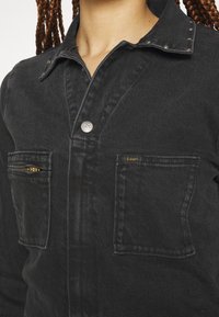 Gros plan d'une veste en jean noire avec deux poches avant, dont une zippée, bouton métallique estampillé Lee et col clouté, portée par une personne aux cheveux tressés.