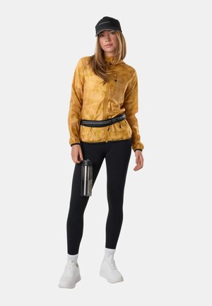 Femme portant une veste moutarde, des leggings noirs, des baskets blanches et une casquette noire, tenant une bouteille d'eau transparente dans une main.