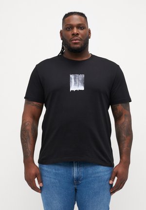 Černé tričko s krátkým rukávem z bavlny s bílým grafickým designem umístěným uprostřed hrudníku, obsahující text "Calvin Klein Jeans".