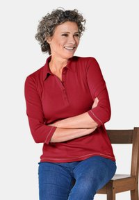 Rode poloshirt met driekwartmouwen, knoopsluiting en contrasterende stiksels. De stof heeft een gladde textuur en een getailleerd ontwerp.