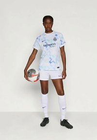 Ανοιχτό μπλε μοτίβο ποδοσφαιρικής φανέλας με λευκά σορτς. Ο παίκτης κρατά μια μπάλα ποδοσφαίρου, φοράει μαύρα παπούτσια. Το λογότυπο Nike είναι ορατό στη φανέλα και στα παπούτσια.