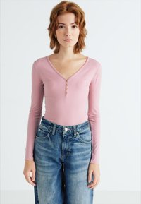 Fittet ribbestrikket langermet topp i lys rosa med dyp V-hals og tre-knappers placket; kombinert med høydevidde blå denimjeans.