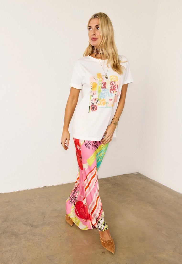 Ξανθιά γυναίκα που φοράει πολύχρωμο παντελόνι με αφηρημένο μοτίβο, λευκό γραφικό t-shirt, καφέ μυτερά παπούτσια και χρυσά αξεσουάρ, στέκεται μπροστά σε λευκό απλό τοίχο.