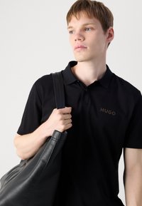 Juodas polo marškinėliai su švelniu tekstūru, puošti rudu logotipu ant krūtinės. Modelis laiko didelę juodą odinę rankinę su pečių diržais.