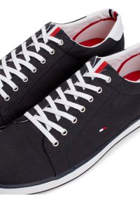 Paire de baskets basses en toile noire avec lacets blancs, semelles blanches, détails rayés rouges et bleu marine, et petit logo drapeau sur le côté.