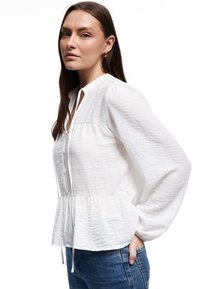 Hvid langærmet skjorte i let stof, med peplum afslutning, knapfront og blød tekstur med let rynkeeffekt.