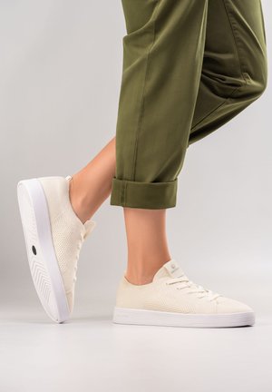 Helle beigen Sneaker aus atmungsaktivem Material mit strukturiertem Obermaterial, weißer Sohle und flachen Schnürsenkeln, kombiniert mit aufgerollten olivgrünen Hosen.