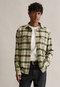 Groen-wit geruit flanellen overhemd met twee borstzakken, knoopsluiting aan de voorkant en lange mouwen; gedragen over een wit shirt en zwarte jeans.