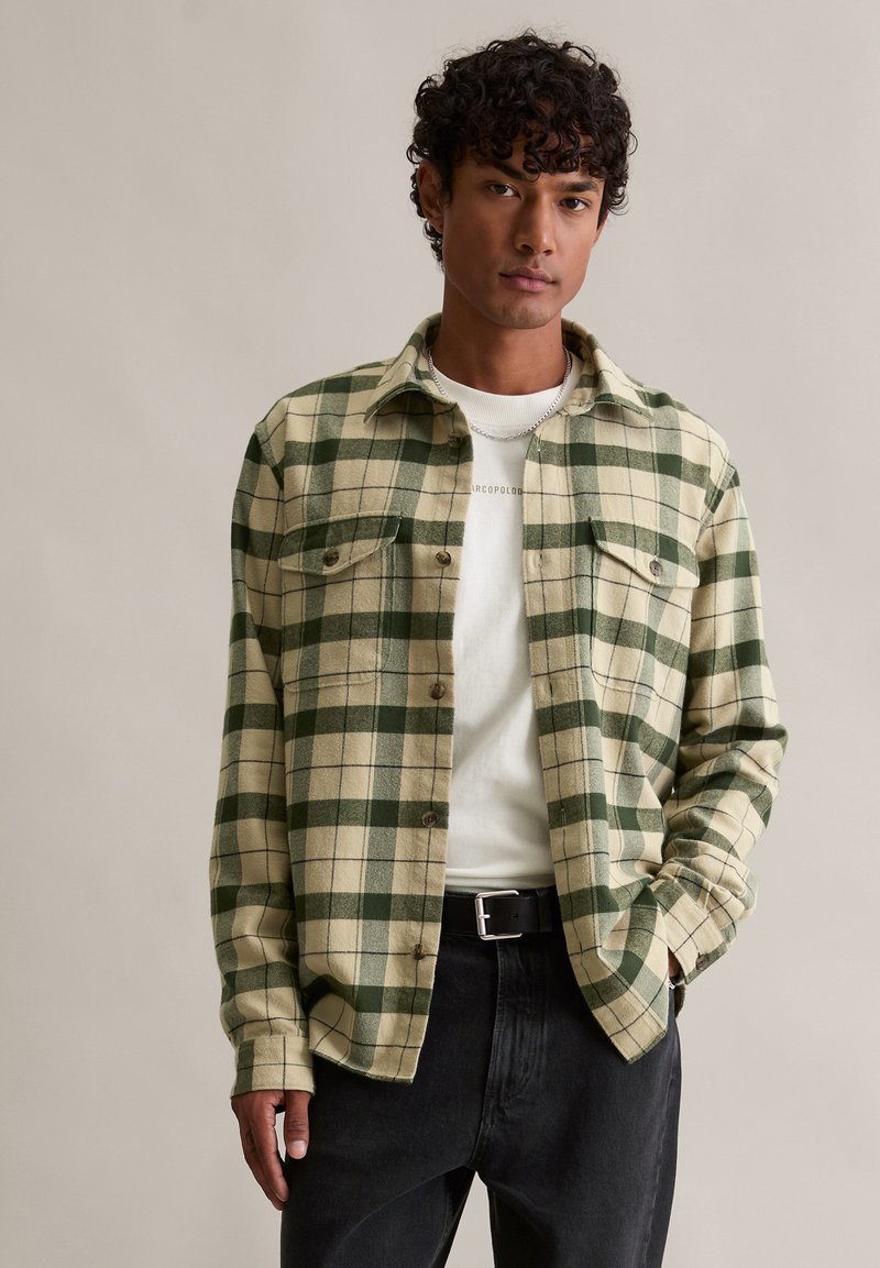 Groen-wit geruit flanellen overhemd met twee borstzakken, knoopsluiting aan de voorkant en lange mouwen; gedragen over een wit shirt en zwarte jeans.