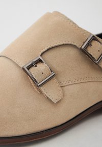 Beige Wildleder Slipper mit glatter Textur, doppelten Metall-Schnallenakzenten und einer leicht spitzen Zehenpartie.