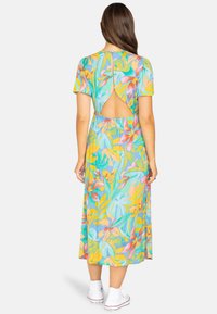 Fließendes Midi-Kleid in Hellblau, mit kurzen Ärmeln, einem Rückenausschnitt und einem elastischen Taillenbund, verziert mit bunten Blättern. Weiße Sneakers.
