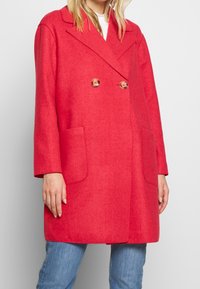 Mujer vistiendo un abrigo rojo brillante de doble botonadura con grandes botones, bolsillos de parche y jeans azules, de pie contra un fondo plano.