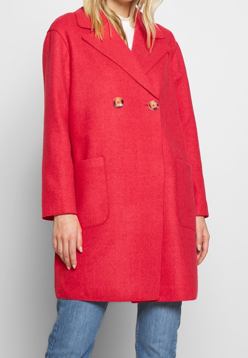 Mujer vistiendo un abrigo rojo brillante de doble botonadura con grandes botones, bolsillos de parche y jeans azules, de pie contra un fondo plano.