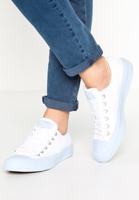 Baskets en toile blanche avec semelles et accents en caoutchouc bleu clair. Caractérisées par un bout arrondi, six œillets et une tige texturée. Portées avec un jean bleu retroussé.