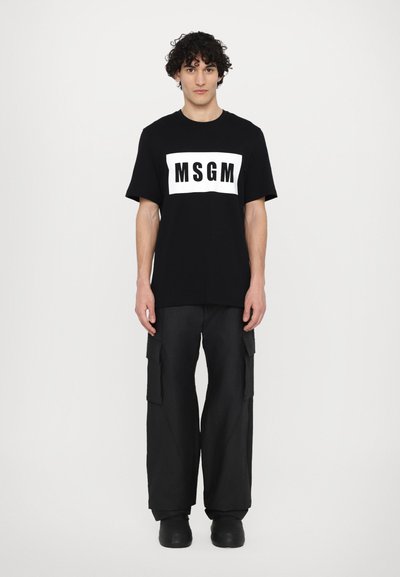 Svart t-shirt med en vit rektangulär logotyp "MSGM" på framsidan. Nederdelen kompletteras med vida svarta cargobyxor och svarta skor.