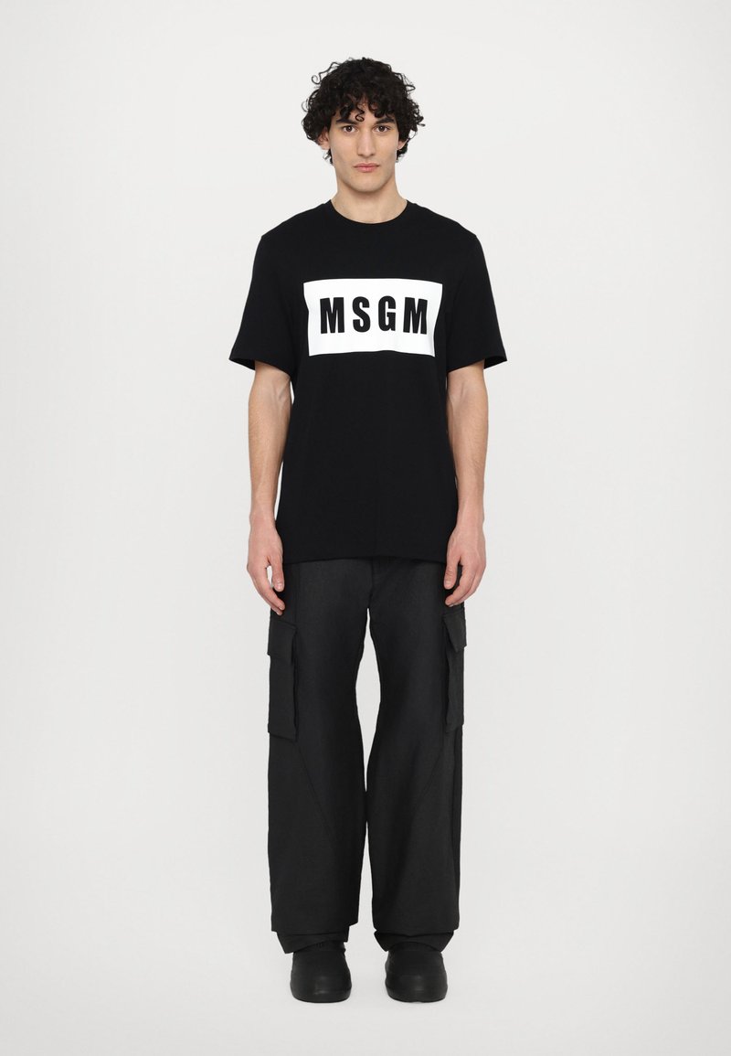 Schwarzes T-Shirt mit einem weißen rechteckigen Logo "MSGM" auf der Vorderseite. Kombiniert mit weit geschnittenen schwarzen Cargo-Hosen und schwarzen Schuhen.