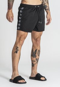 Čierne plavky s elastickým pásom a šnúrkou, s bielym vzorovaným akcentom pozdĺž boku. Nosené s čiernymi šľapkami.