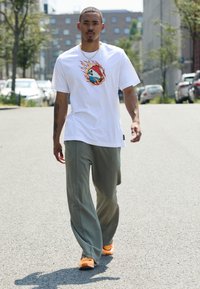 Bărbat care merge pe stradă îmbrăcat cu un tricou alb cu grafică de casă în flăcări, pantaloni verzi, adidași portocalii și lănțișor de aur.