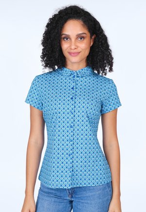 Button-up shirt in blue fabric featuring a wave pattern, short sleeves, a collar, and a fitted design. Paired with blue jeans.