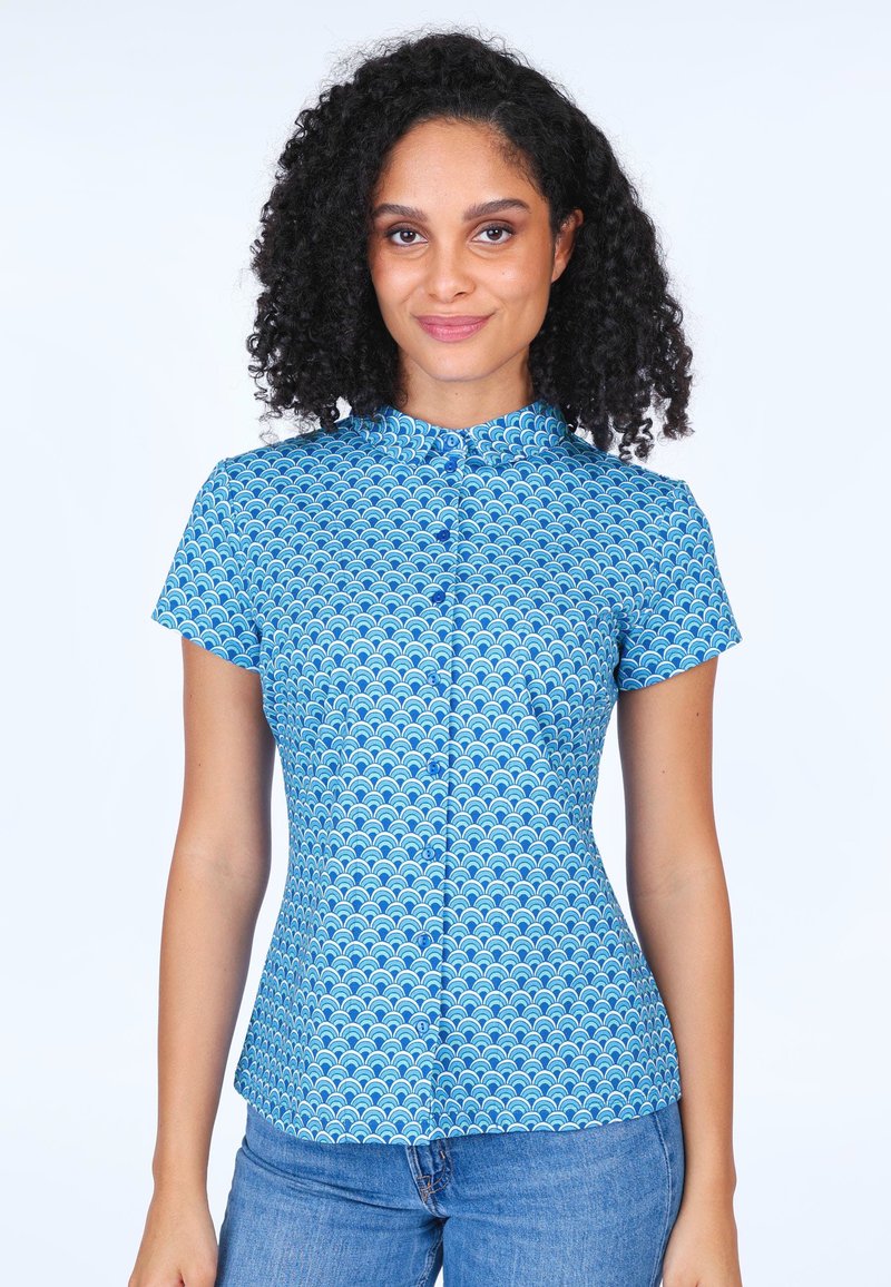 Button-up shirt in blue fabric featuring a wave pattern, short sleeves, a collar, and a fitted design. Paired with blue jeans.