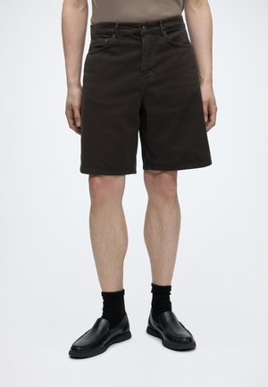Muž oblečený v čiernych rifľových šortkách po kolená, s čiernymi ponožkami a čiernymi slip-on topánkami, stojaci na jednoduchom bielom pozadí.
