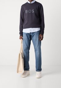 Marinblå crewneck sweatshirt med "BOSS" logotyp, ljusblå skjorta under, blå jeans, vita sneakers och en beige tote-väska.