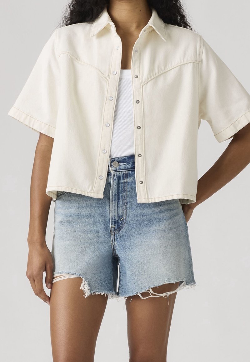 Personne portant une chemise crème à manches courtes boutonnée sur un débardeur blanc et un short en jean bleu clair déchiré.