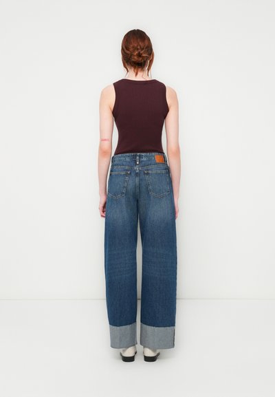 Débardeur bordeaux associé à un jean bleu taille haute et à jambes larges, avec des ourlets retroussés et une pièce en cuir marron à l'arrière.