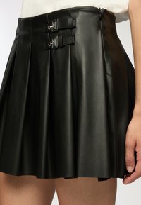 Gonna plissettata in pelle nera con due dettagli a fibbia in vita e una texture liscia, che copre parzialmente la parte superiore della coscia.