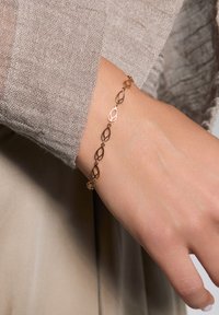 Różowe złote bransoletka z połączonymi owalnymi kształtami, małymi koralikami i zapięciem. Na tle teksturowanej beżowej tkaniny.