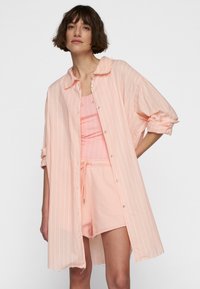 Camicia oversized rosa chiaro con bottoni, decorata con strisce verticali bianche, abbinata a una canotta e pantaloni corti dello stesso colore. Tessuto morbido con maniche arrotolate.