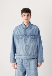 Denim jacket in light blue, featuring a button front, two chest pockets, and standard collar. Fabric has a slightly worn texture.