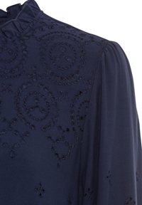 Blouse bleu marine ornée de motifs brodés complexes et de détails découpés, avec un col à volants et des manches délicatement bouffantes.