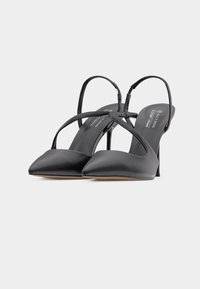 Μαύρες γόβες με μυτερή μύτη, λου ράκια που διασταυρώνονται και τακούνια στιλέτο σε ανοιχτό γκρι φόντο.