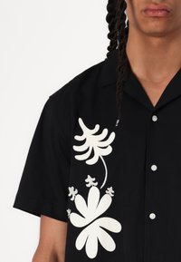 Korte mouw zwarte shirt met grote witte bloemembroidery, ronde kraag en knoopsluiting. De stof lijkt glad te zijn met een relaxte pasvorm.