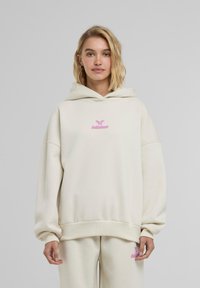 Valkoinen beige oversized huppari, joka on valmistettu pehmeästä puuvillasta. Hupparissa on vaaleanpunainen brodeerattu logo rinnassa ja resorihihansuut. Yksinkertainen muotoilu.