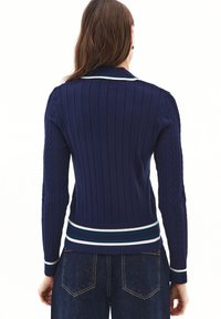 Suéter de punto azul marino con textura acanalada, que presenta mangas de punto trenzado y un dobladillo a rayas en verde, azul y blanco.