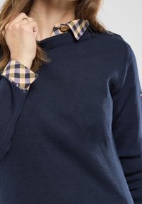 Femme portant un pull bleu marine par-dessus une chemise à carreaux jaunes et bleus, tenant le col du pull, montrant les poignets et le col de la chemise.