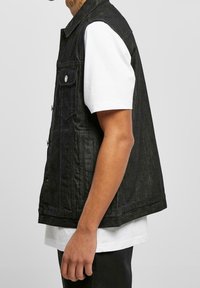 Svart denimväst med knäppning fram, två bröstfickor och en avslappnad passform, bärs över en vit t-shirt. Slät textur och minimal dekoration.