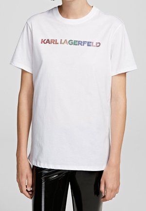 Person trägt ein weißes T-Shirt mit buntem "KARL LAGERFELD" Text auf der Vorderseite und glänzende schwarze Hosen, gezeigt vom Hals bis zu den Hüften.