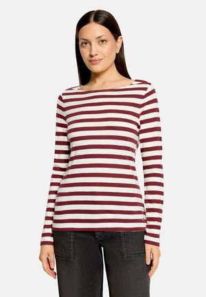 Femme aux cheveux raides et foncés portant une chemise à manches longues blanche et bordeaux rayée, et un jean foncé, debout devant un fond uni.