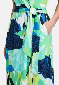 Kolorowa sukienka z dużymi zielonymi i niebieskimi liśćmi, z paskiem w talii i kieszeniami po bokach. Wykonana z lekkiego materiału.