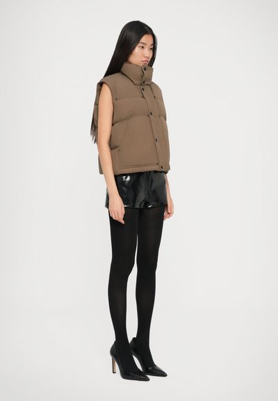 Brun ærmeløs puffervest med høj krave og knaplukning, parret med sorte glossy shorts og sorte strømpebukser i spidse hæle.